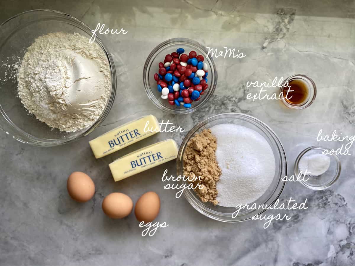 Ingredients on marble counter: flour, sugar, butter, eggs, M&M's, salt, baking soda, vanilla extract.