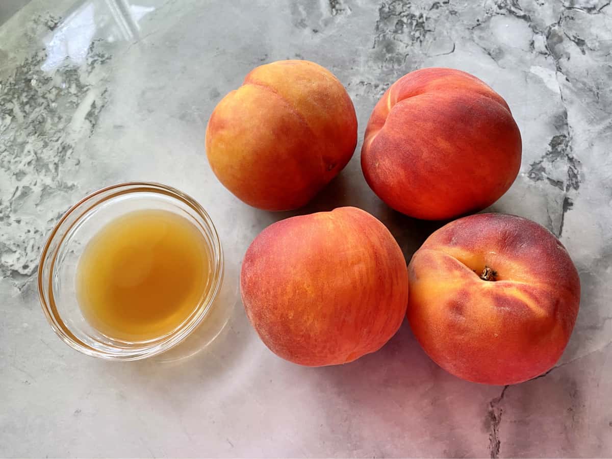 Four peaches and a glass bowl of apple juice on a marble countertop. 
