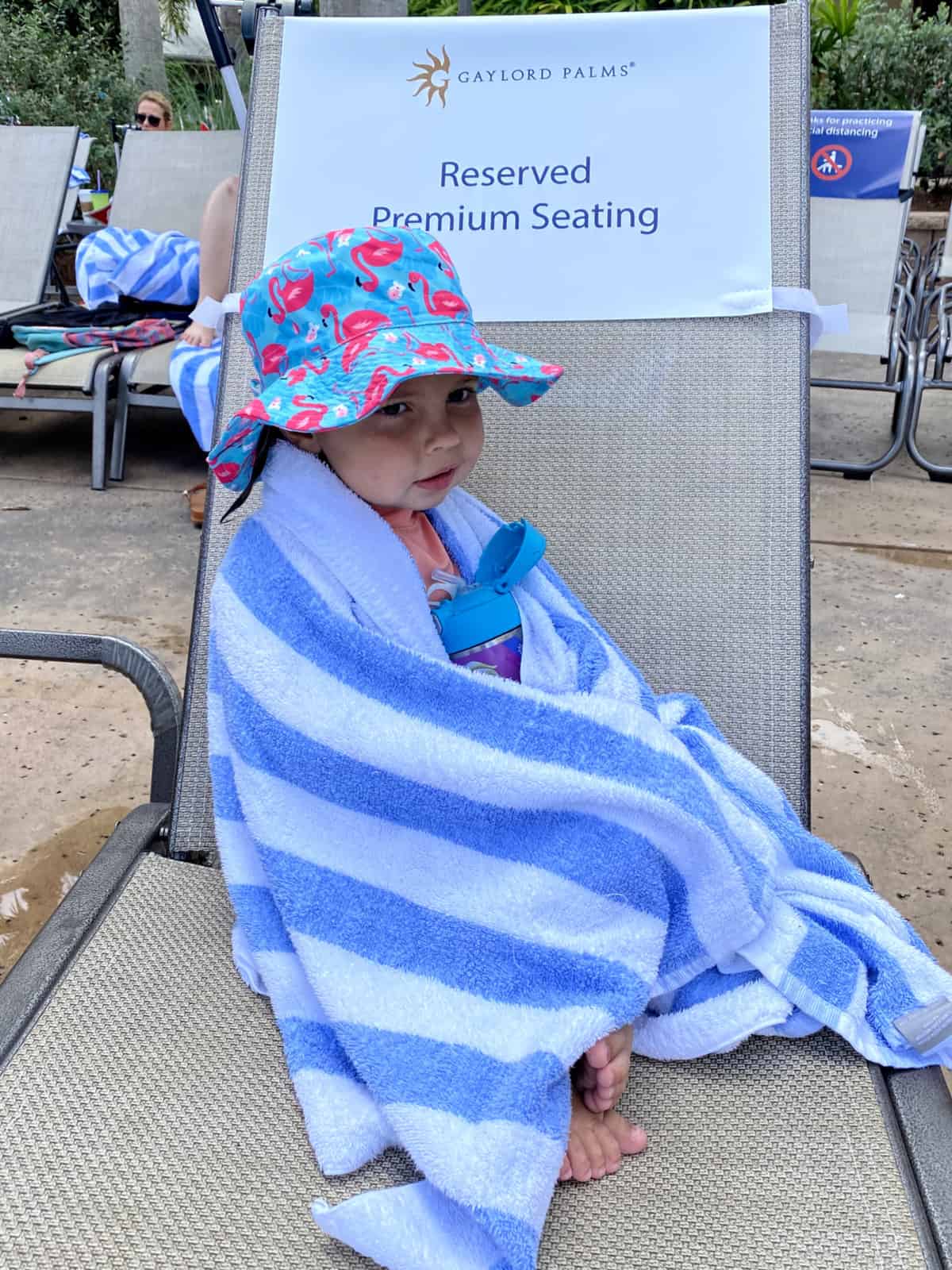 Little girl with flamingo hat wrapped in a white and blue towel sitting on lounge chair.