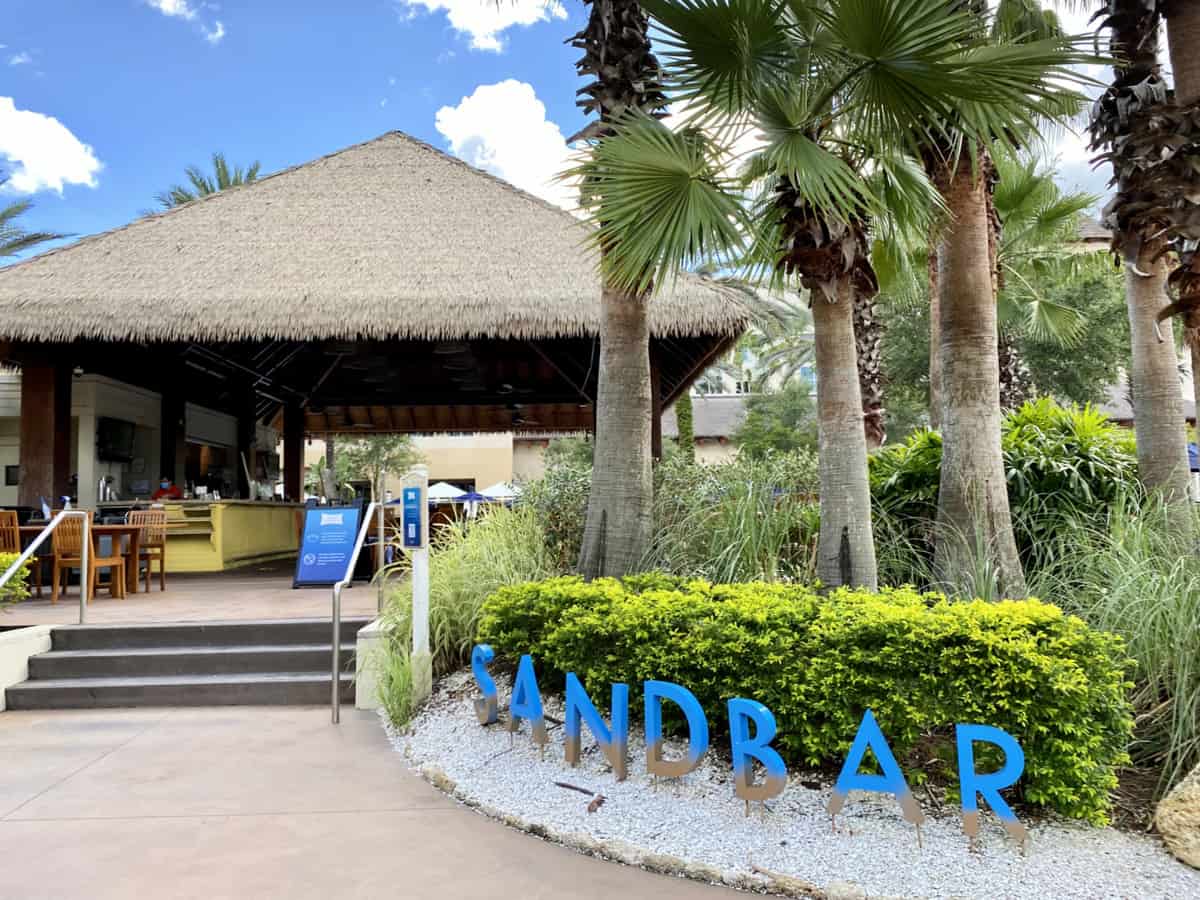 An outside restaurant with palm trees and sign in front of palm bushes that reads "sandbar".