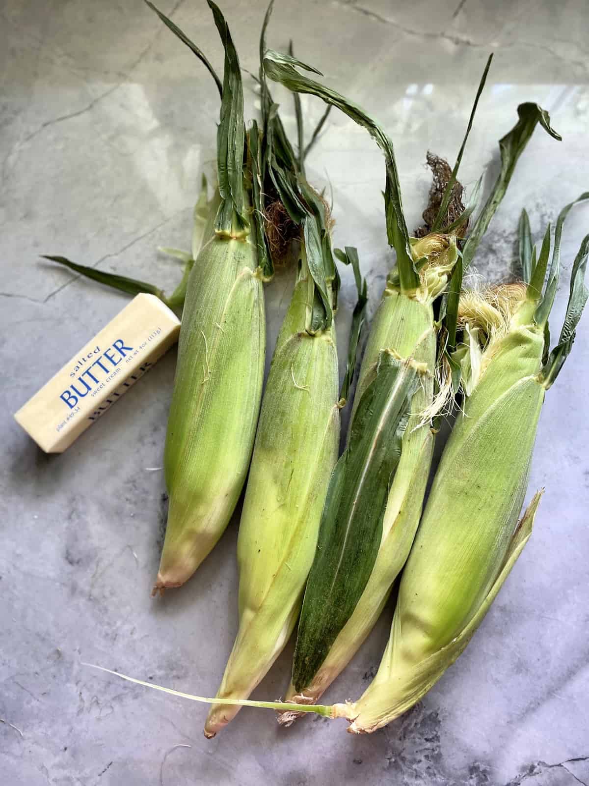 Top view of four ears of corn with a stick of butter on a marble countertop.