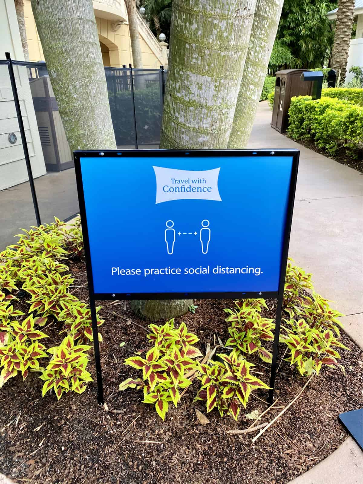 Blue travel with confidence social distancing sign in flower bed in front of trees.