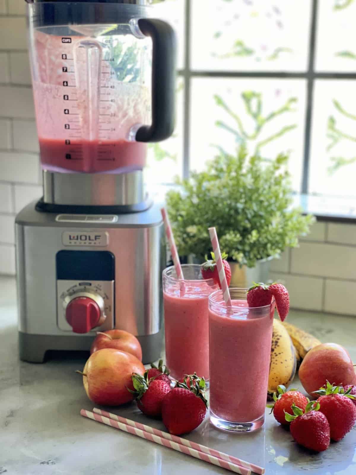 Two glasses with smoothies next to a blender with fruit on the counter. 