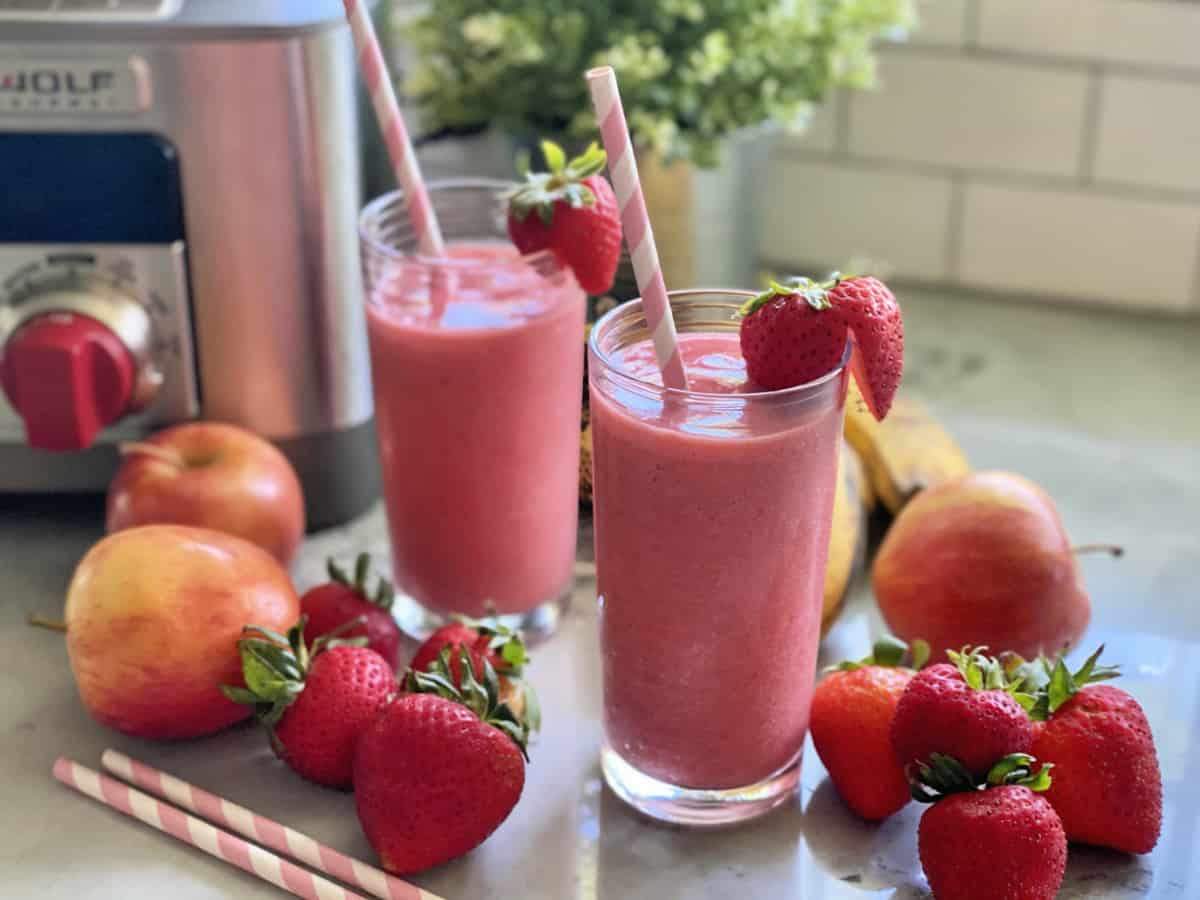 Clear glass with a pink smoothie in it with strawberries on the rim and a paper straw.
