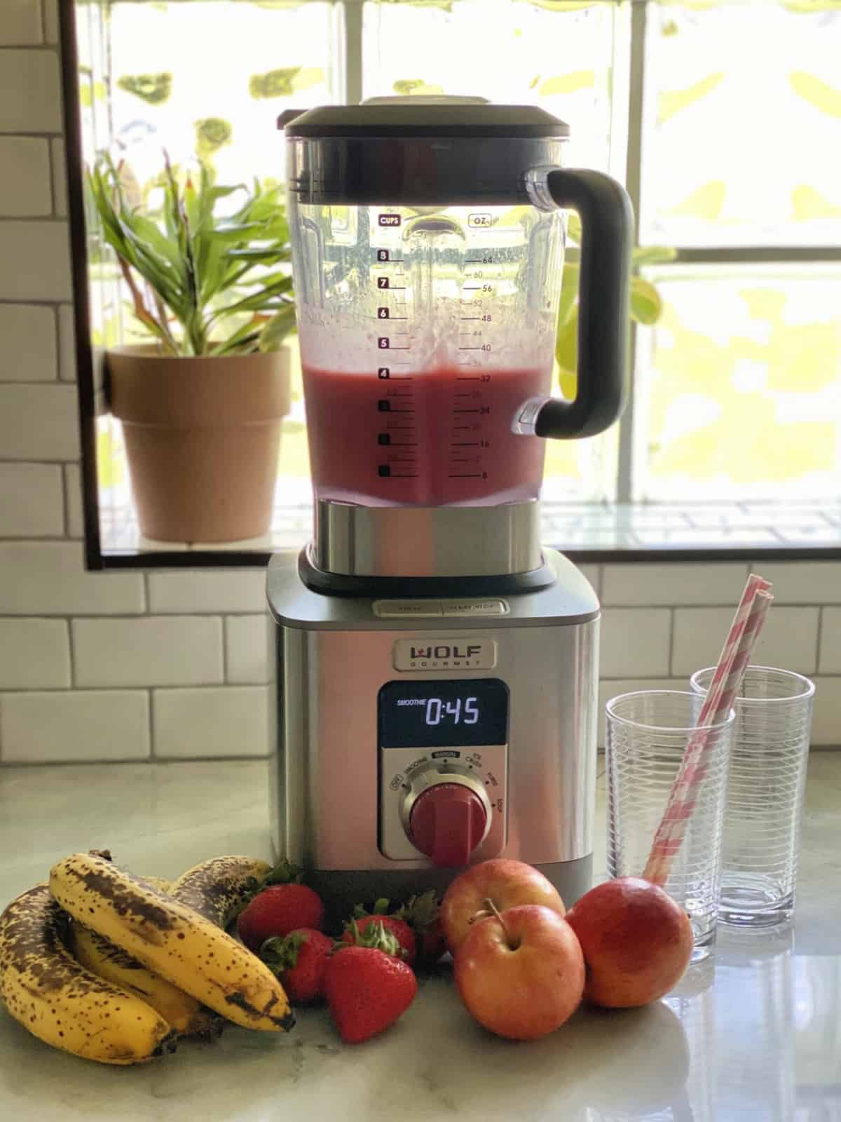 Blender with blended smoothie, fruit and empty glasses on counter around blender.