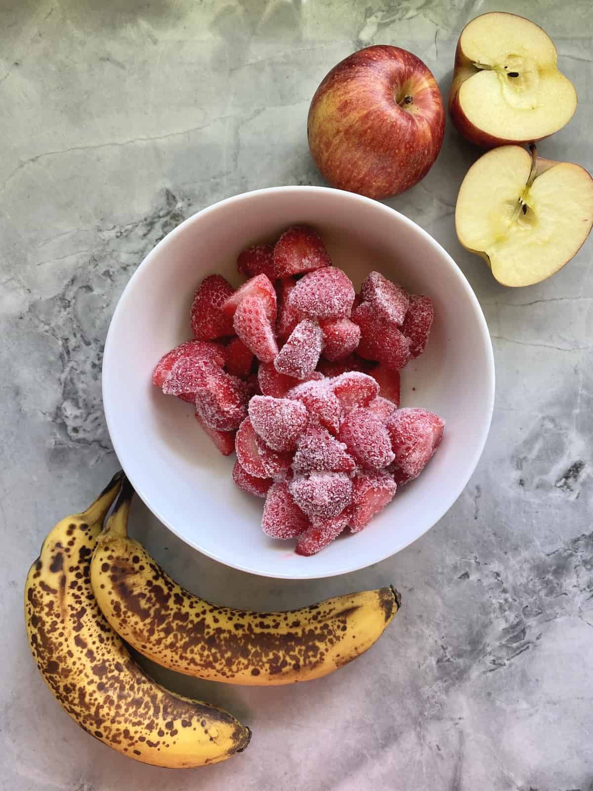 Over ripe bananas, frozen strawberries, cut apples on a marble countertop.