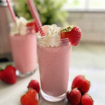 Two pink milkshakes topped with whipped cream and strawberries with straws on a marble counter.