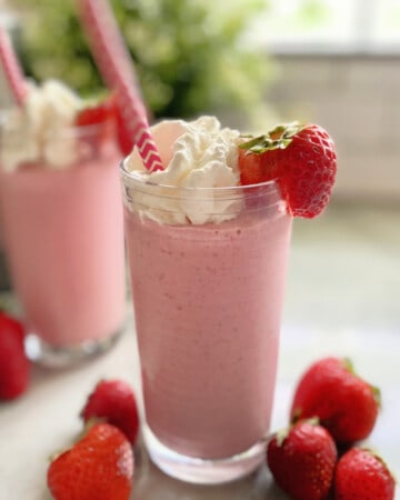 Two pink milkshakes topped with whipped cream and strawberries with straws on a marble counter.