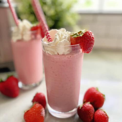 Two pink milkshakes topped with whipped cream and strawberries with straws on a marble counter.