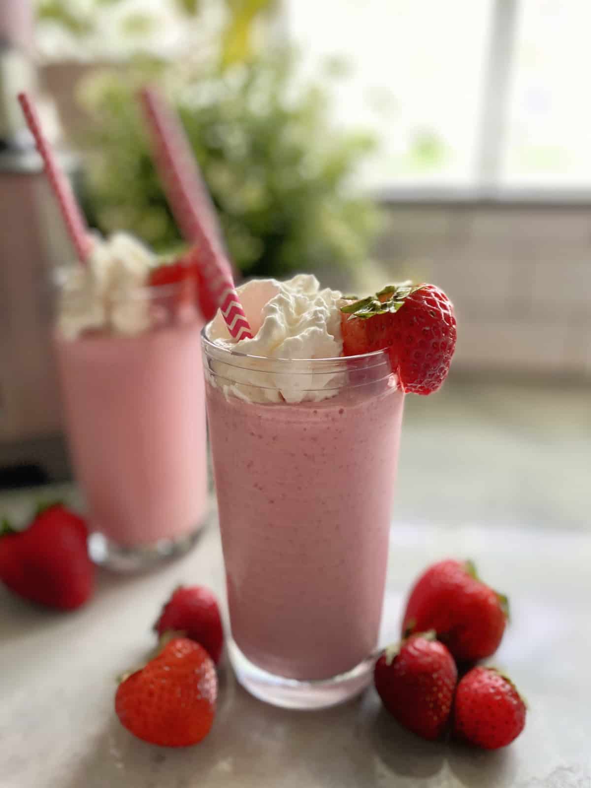 Clear glass of strawberry milkshake with whipped cream and a fresh strawberry on the rim. 