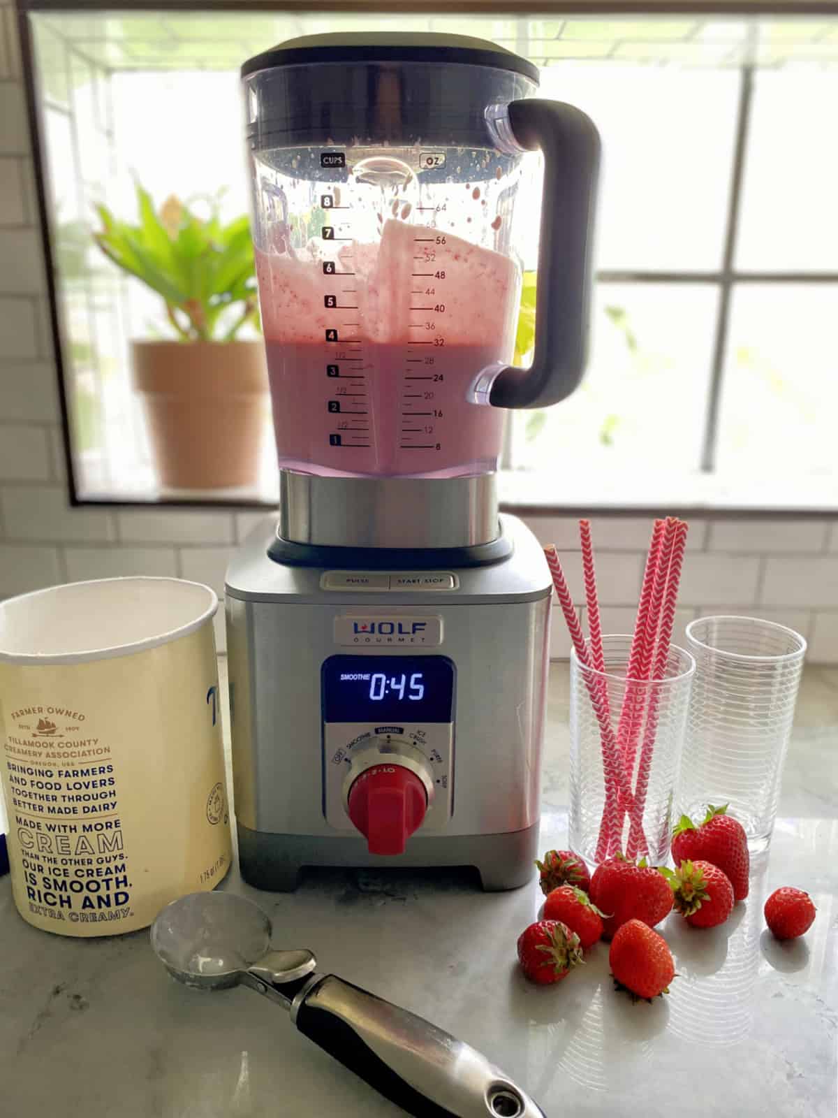 Blender with pink milkshake, empty glasses, and ice cream container with scoop.