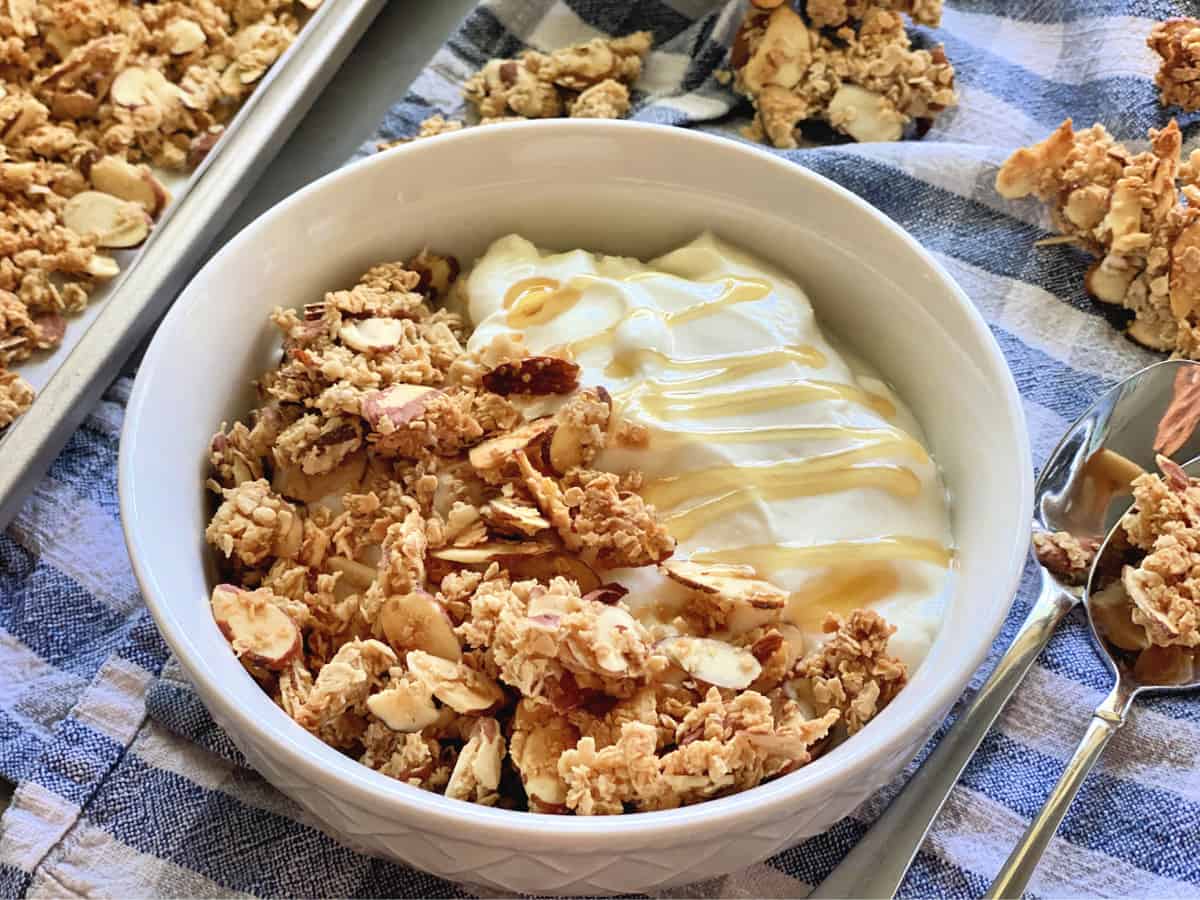 Close up of white bowl with granola, yogurt, and honey on a blue and white checkered cloth.