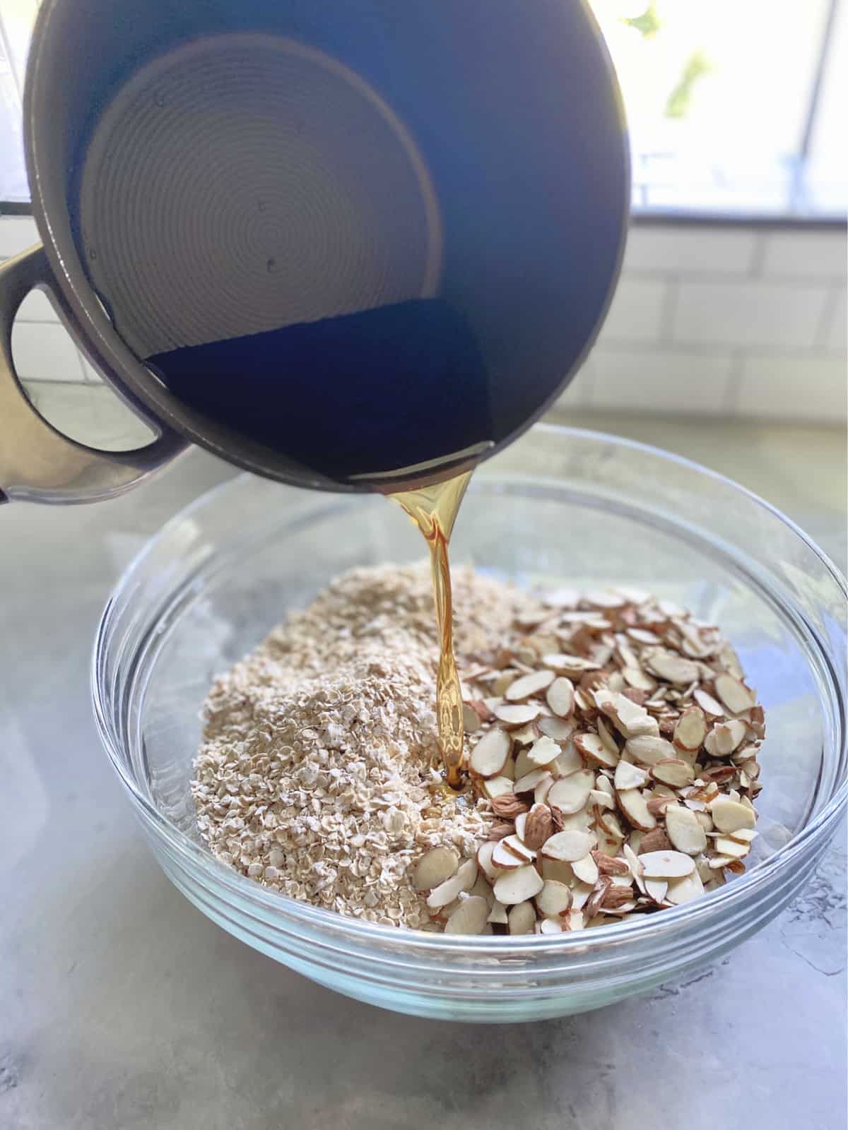 Pot pouring brown liquid into a glass bowl of oats and almond slices on a marble countertop.