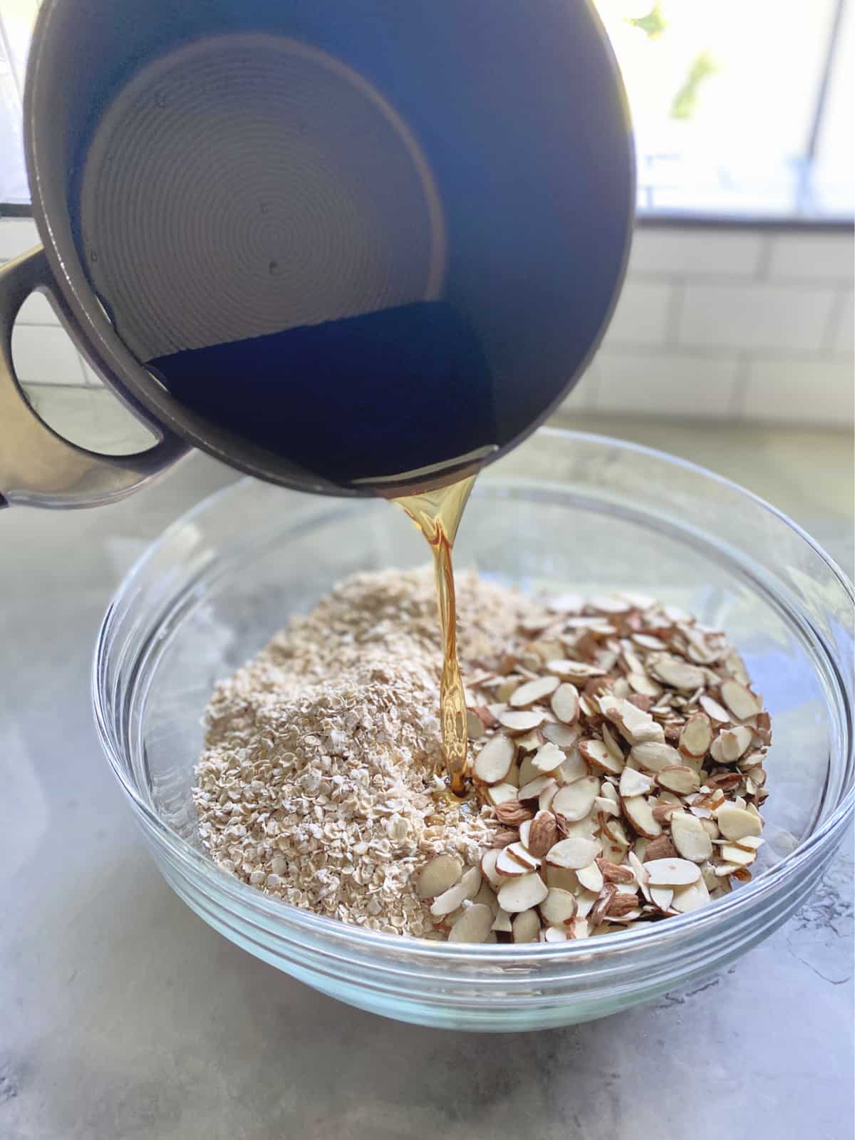 Pot pouring brown liquid into a glass bowl of oats and almond slices on a marble countertop. 