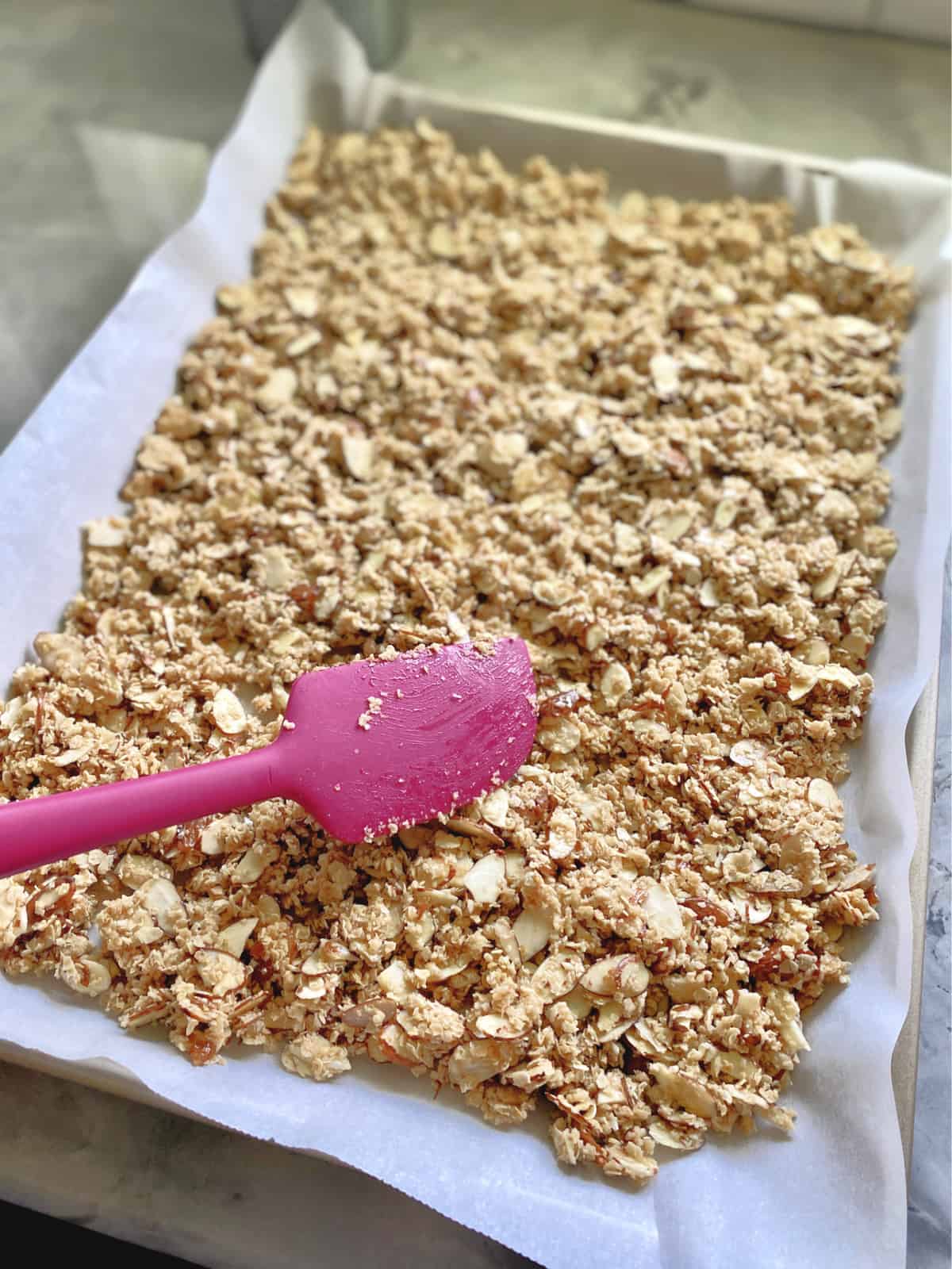 Mixing granola on a parchment lined baking sheet with pink spatula. 
