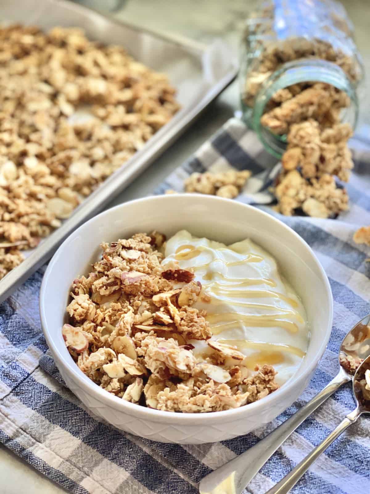 White bowl of yogurt with granola on blue cloth with spoons and granola tray in background. 