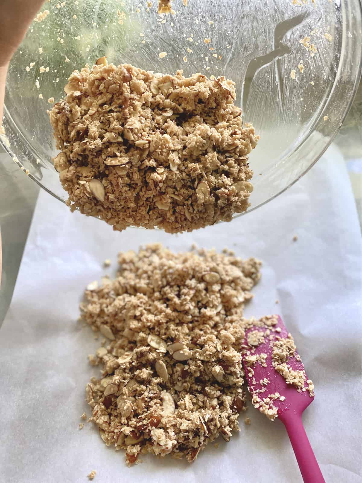 Glass bowl of granola pouring onto a parchment lined baking sheet with pink spatula.