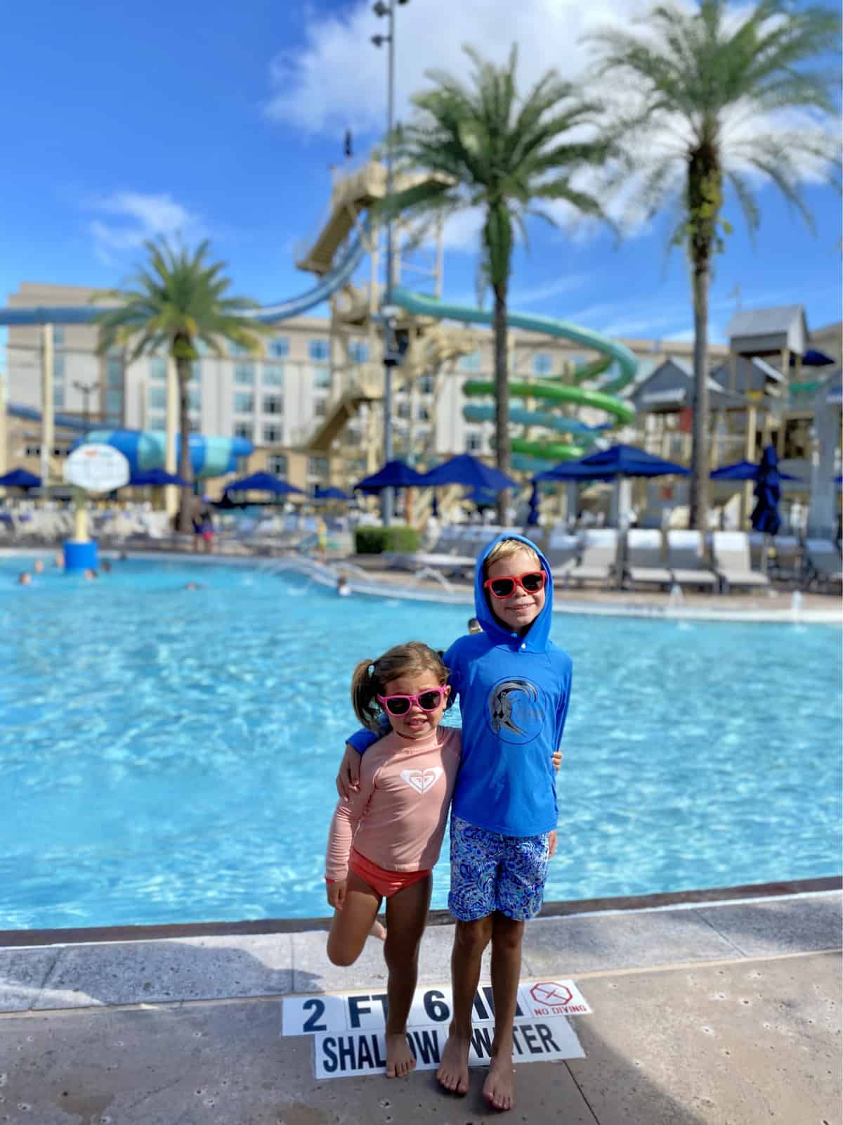 Two children, boy and girl standing in front of a pool with water slides in the background.
