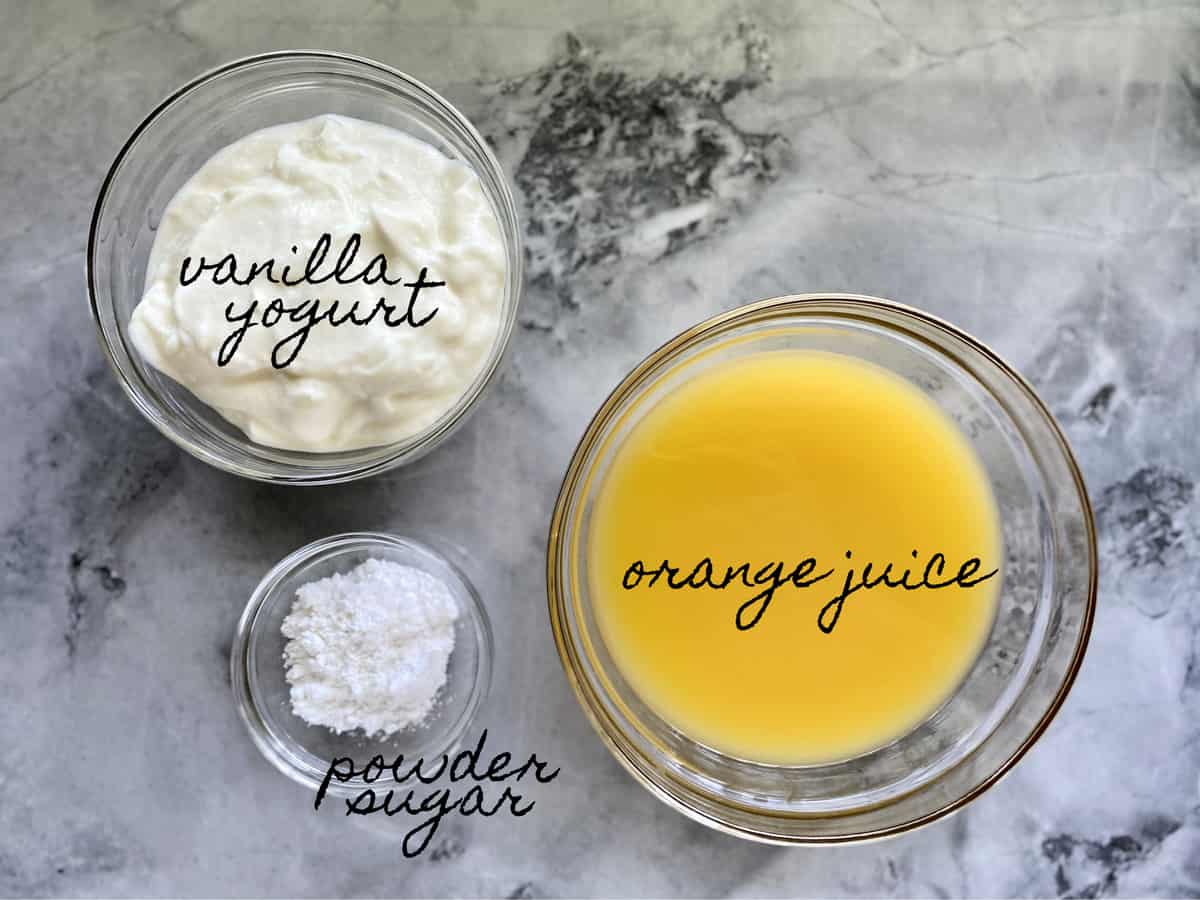 Ingredients on marble counter in glass bowls; vanilla yogurt, powder sugar, and orange juice. 