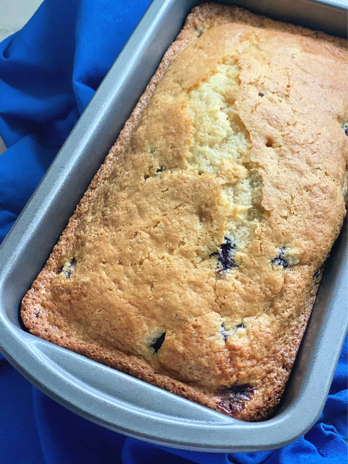 Baked blueberry bread in loaf pan resting on a blue cloth.