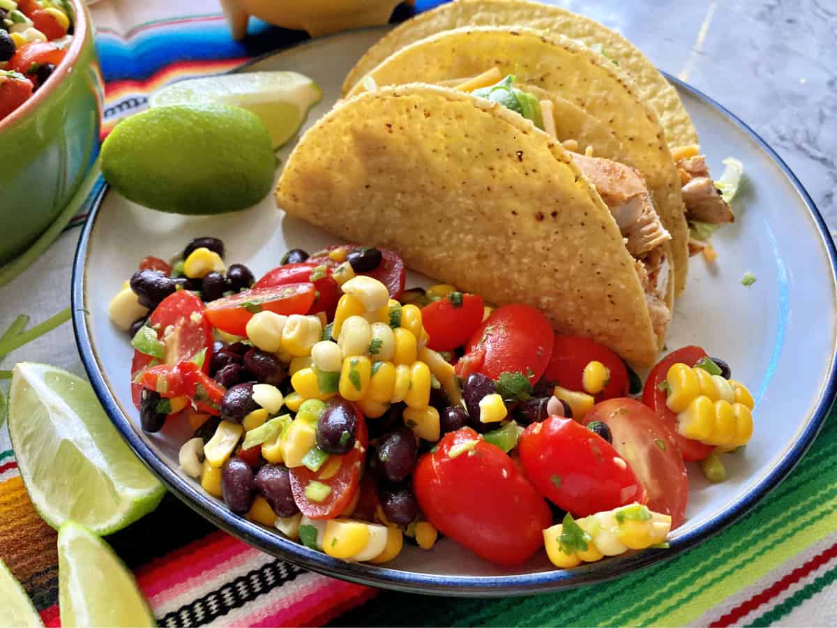 White plate with corn, black beans, and tomatoes next to chicken tacos and lime. 