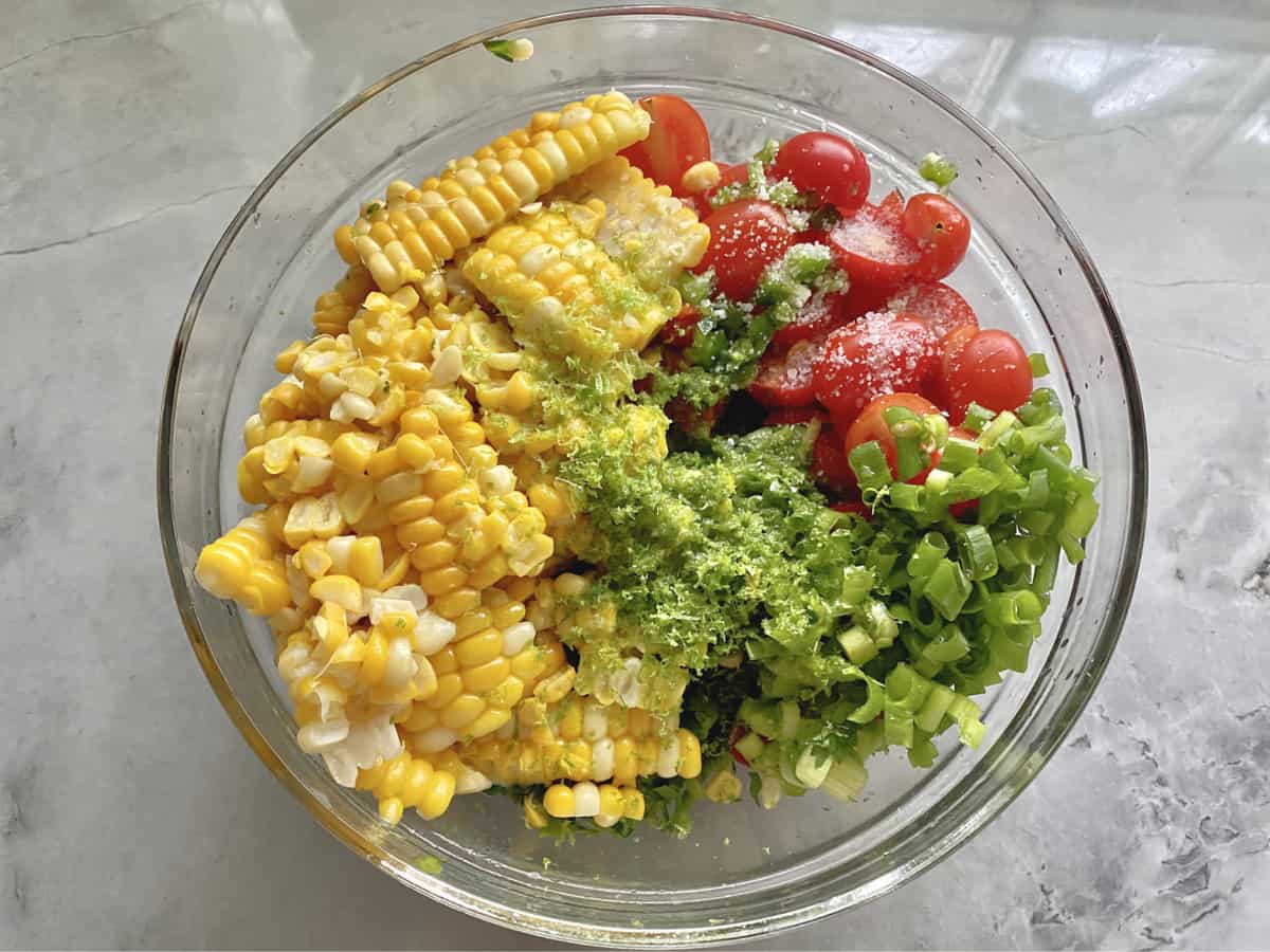 Top view of a glass bowl with corn kernels, sliced tomatoes, salt, green onions, and lime zest.