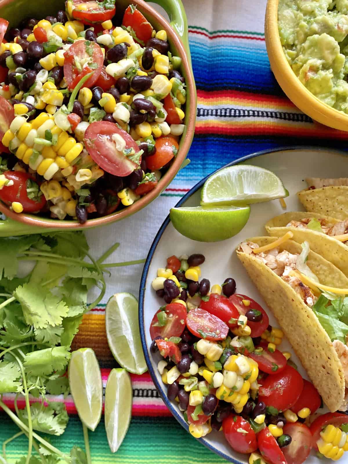 Top view of corn, black beans, and tomatoes on a plate with tacos and lime. 