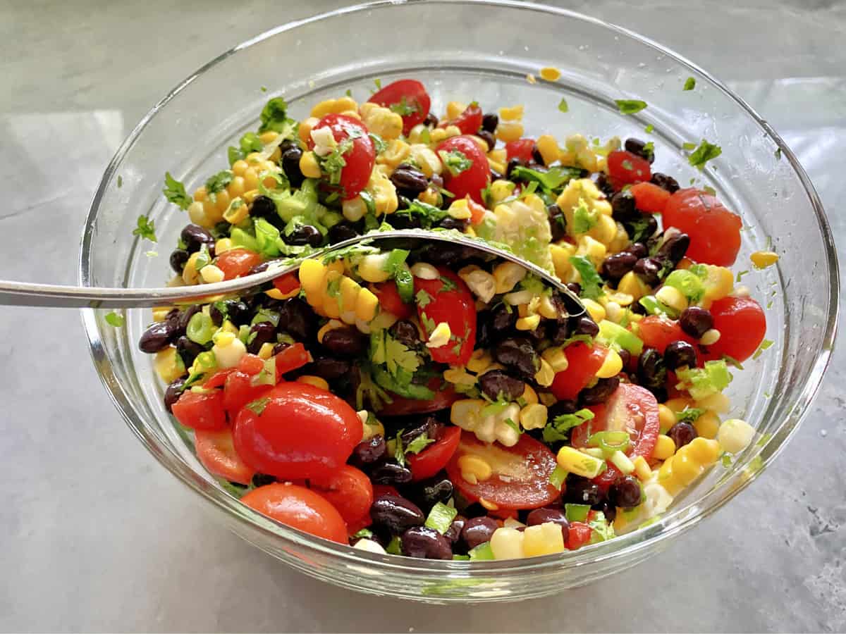 Glass bowl with black beans, sliced grape tomatoes, and corn with spoon mixing.