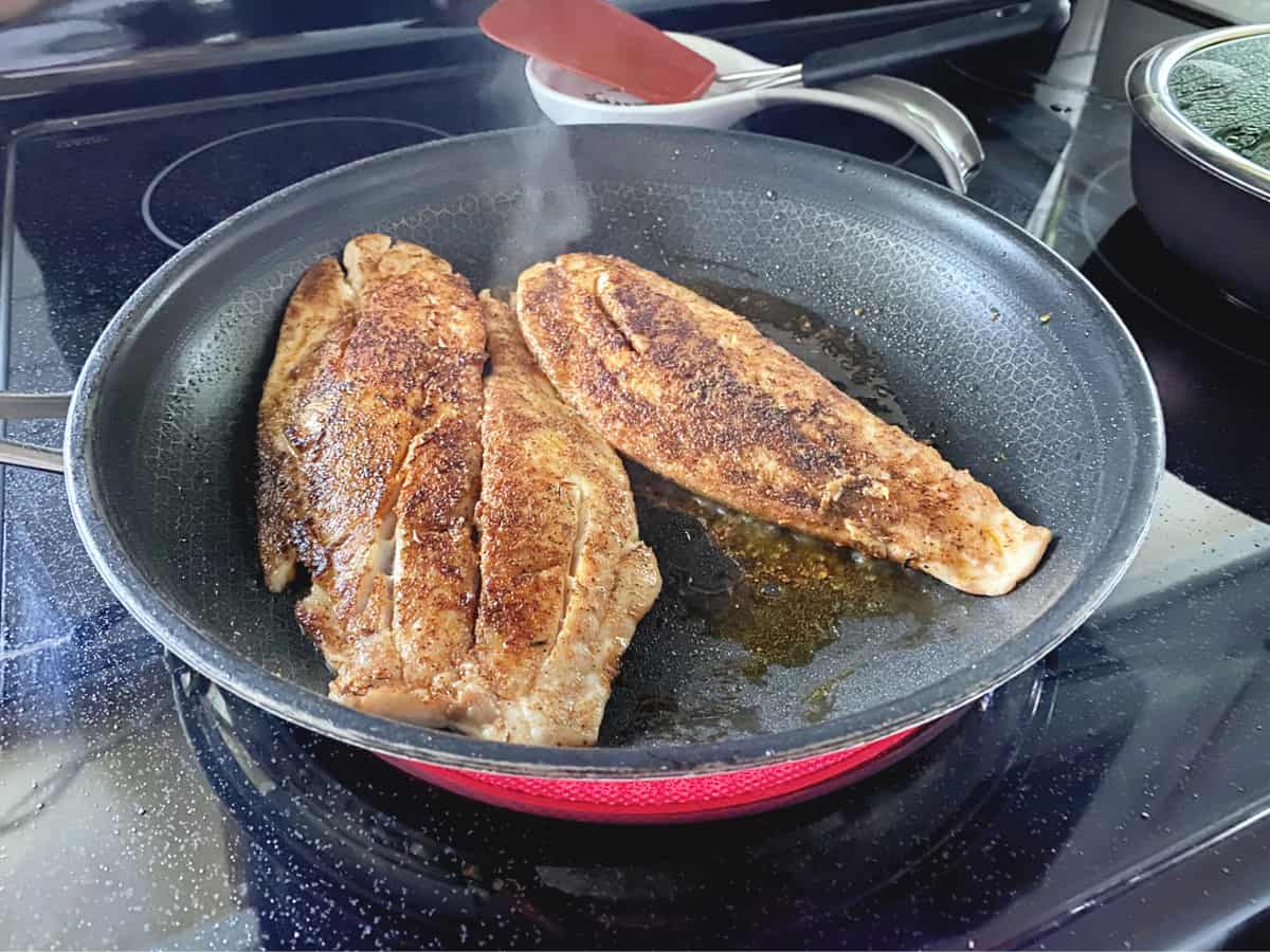 Black frying pan on stove top with seasoned fish frying in the pan.