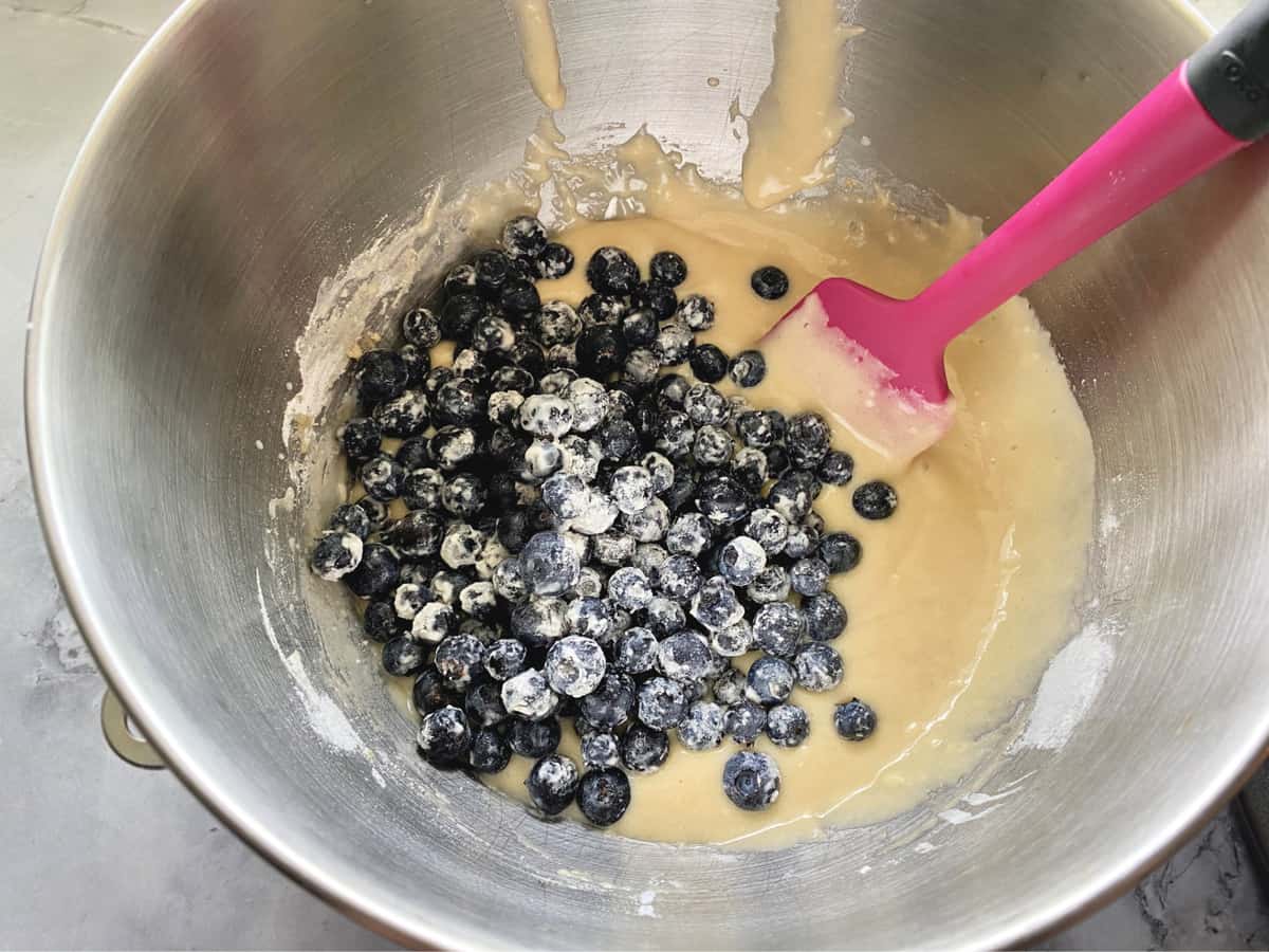 Batter with flour blueberries in a silver bowl with a pink and black spatula. 
