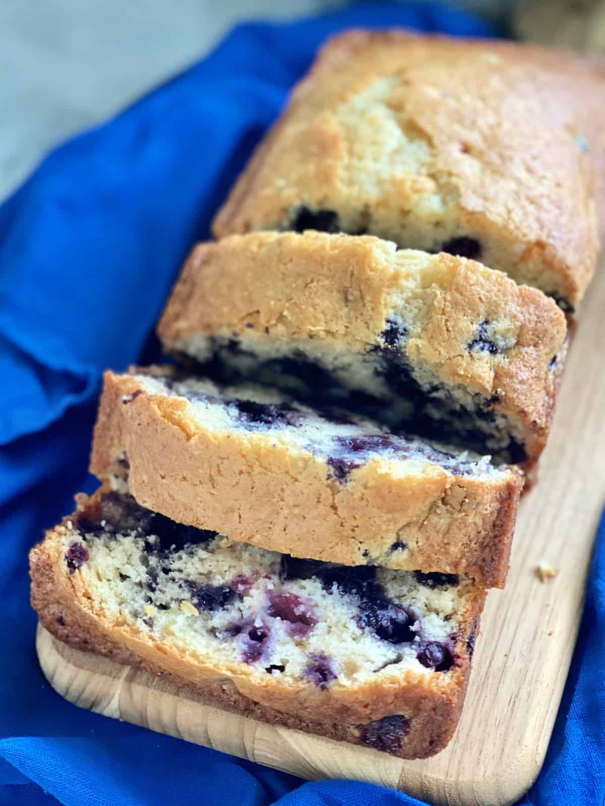 Three slices of blueberry bread on  a wood cutting board with blue fabric. 