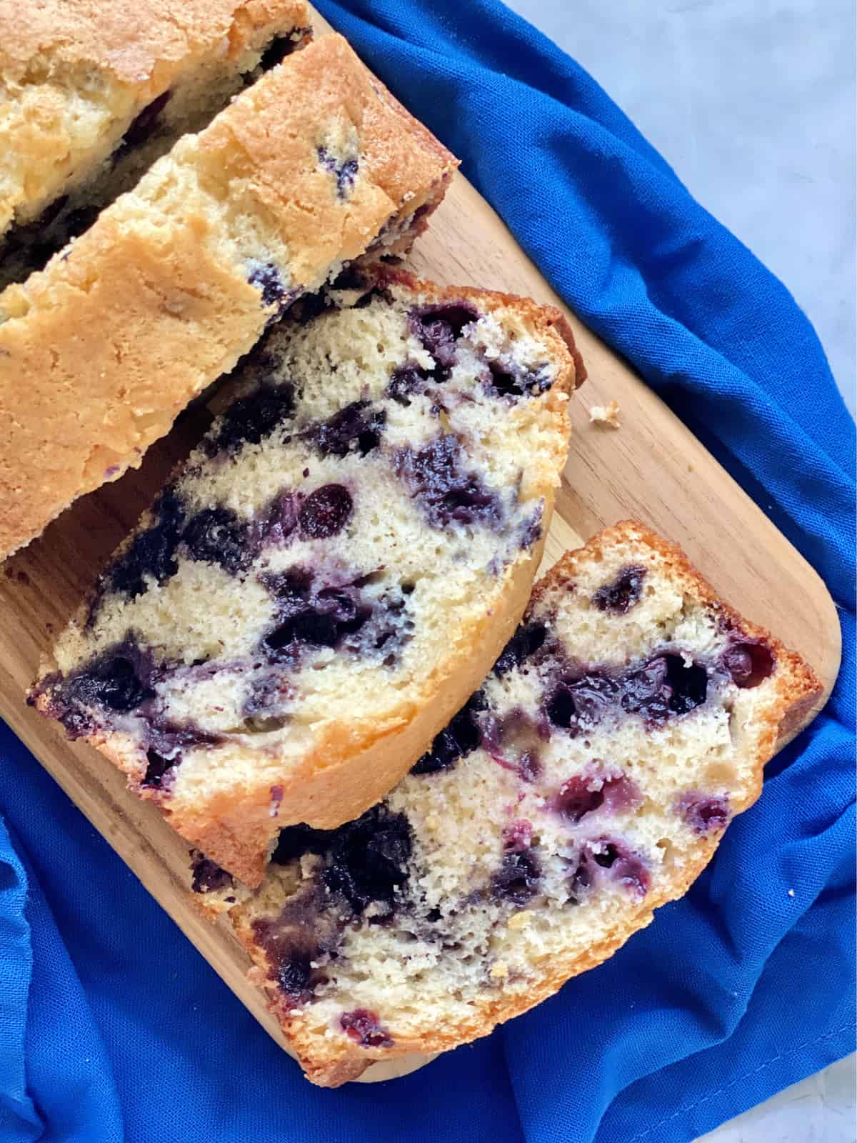 Top view of three slices of blueberry bread on a wood cutting board on a blue cloth.