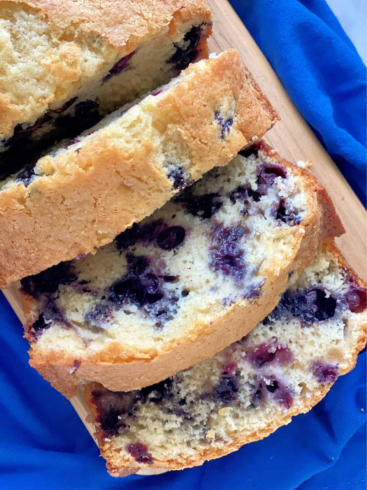 Top view of three slices of blueberry bread sitting on a cutting board and blue cloth.