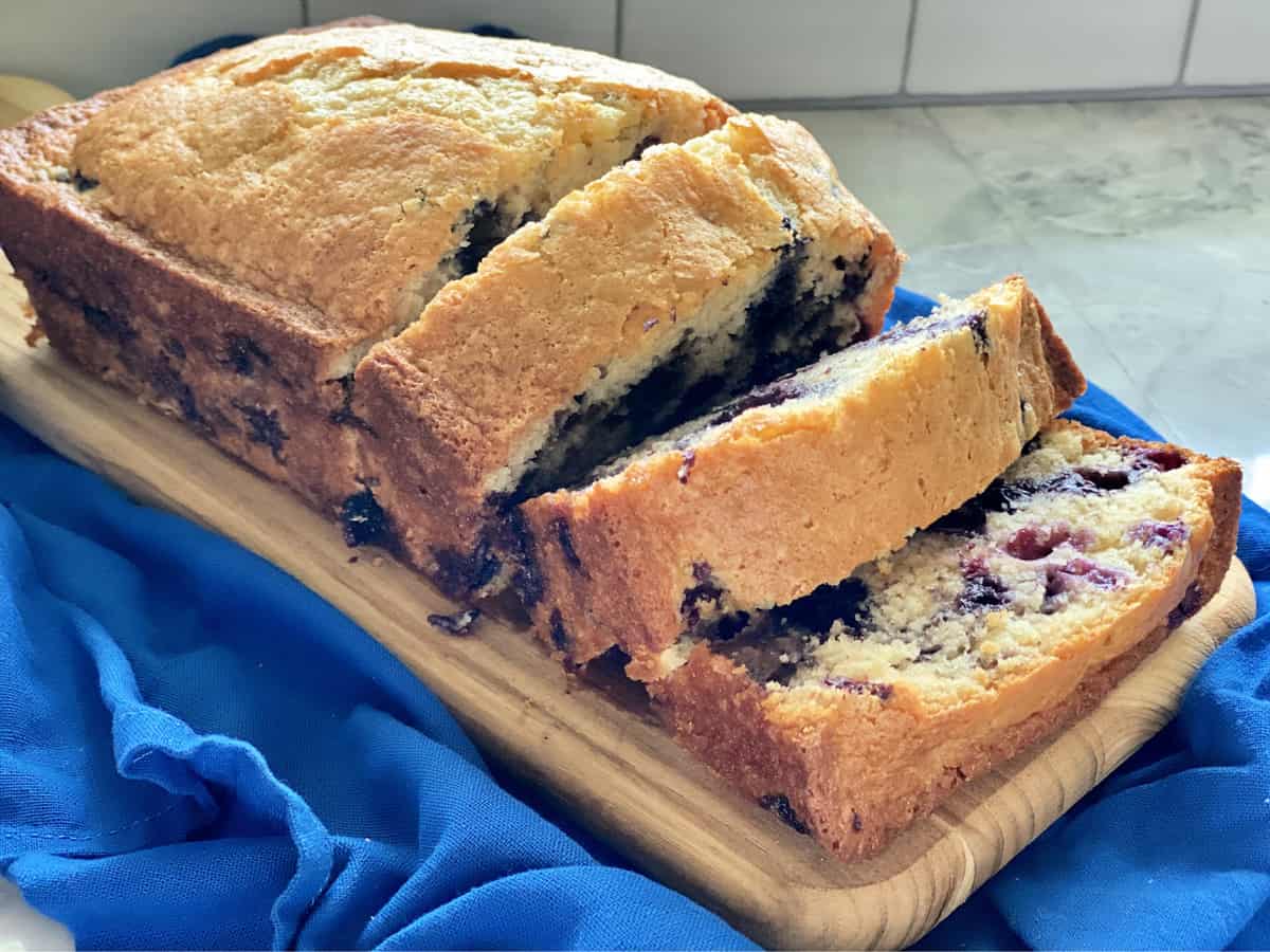 Loaf of bread with blueberries in it with three slices on a wood cutting board. 