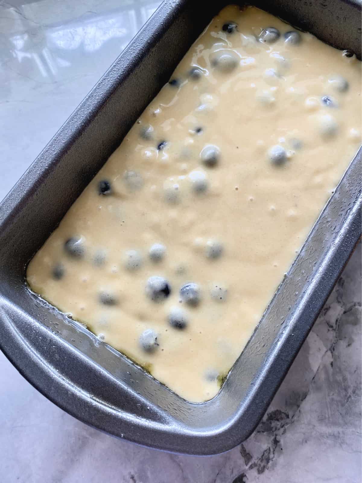 Cake batter with blueberries in a silver loaf pan on marble countertop.