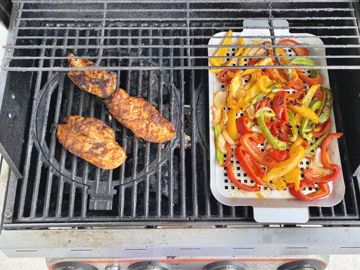 Chicken on a grill next to a grill pan full of multi-color bell peppers and onion.