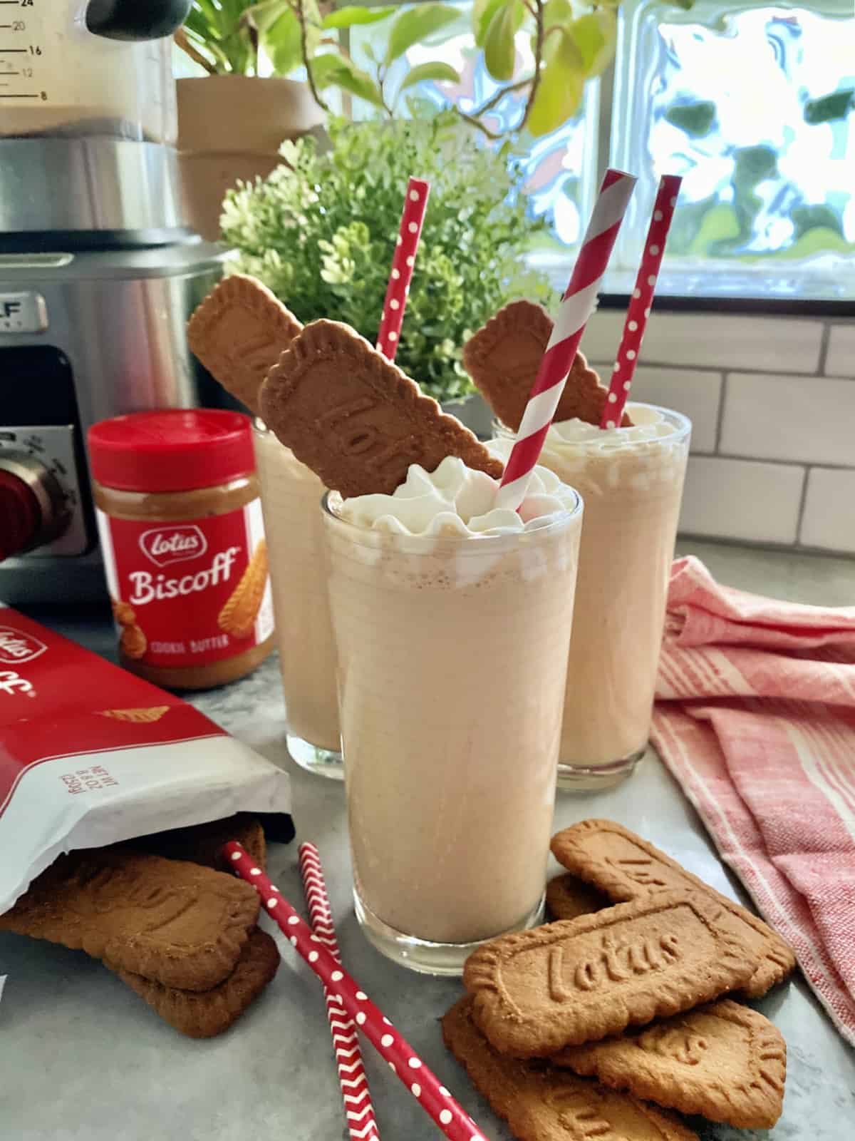 Three clear glasses with a light brown milkshake in it with whipped cream, red straw, and lotus cookie.