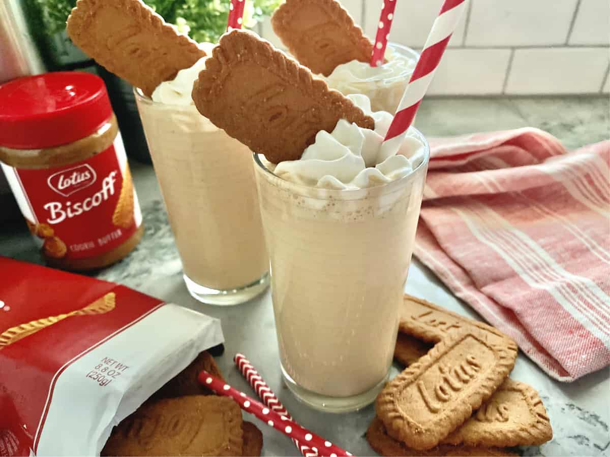 Lotus cookies on counter with three glasses full of a tan milkshake with whipped cream.