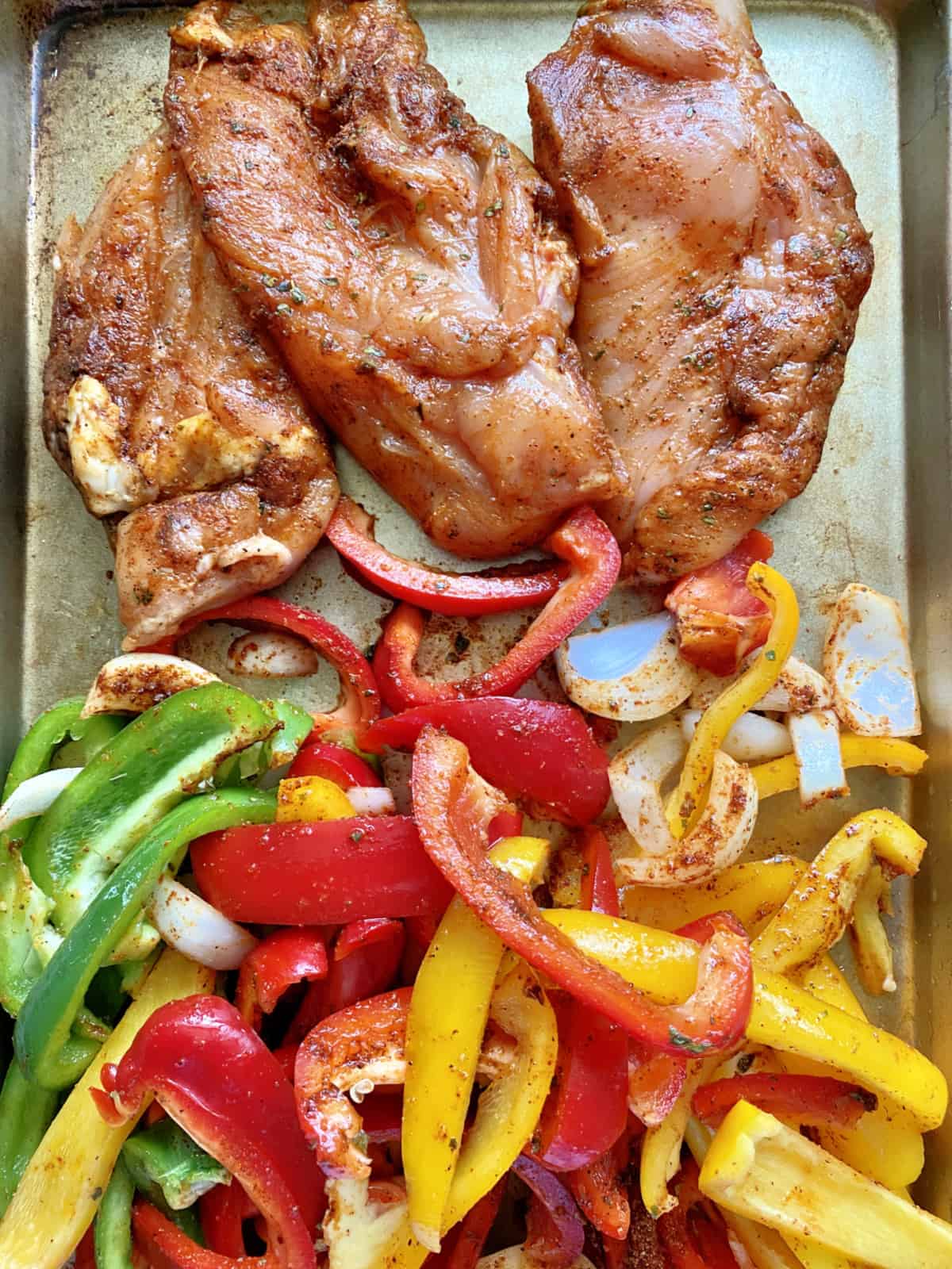 Marinaded chicken breast with mixed color bell peppers and onions on a baking sheet. 