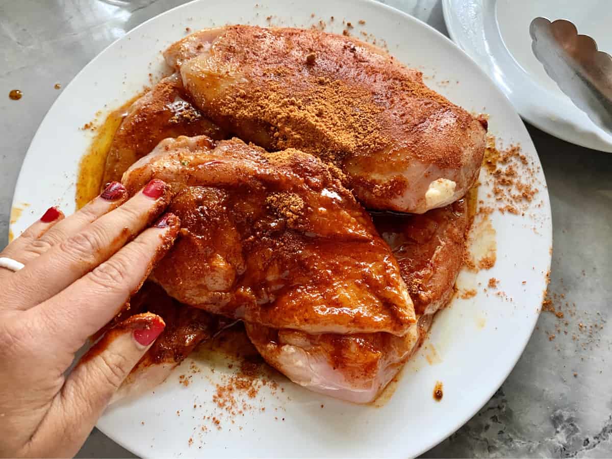 Hand with red nail polish massage red seasoning on chicken breast on white plate. 