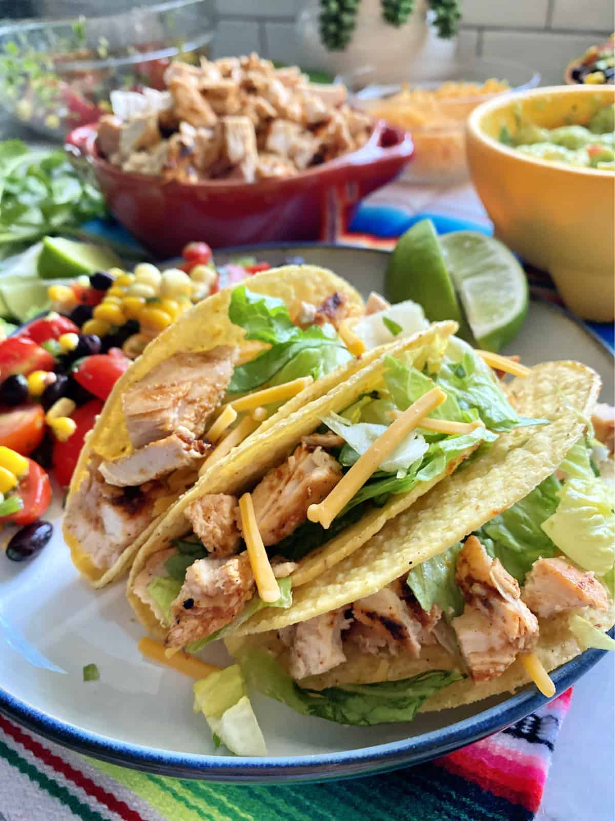 Three hard taco shells with chicken, cheese, and lettuce on a plate with Mexican food around it.