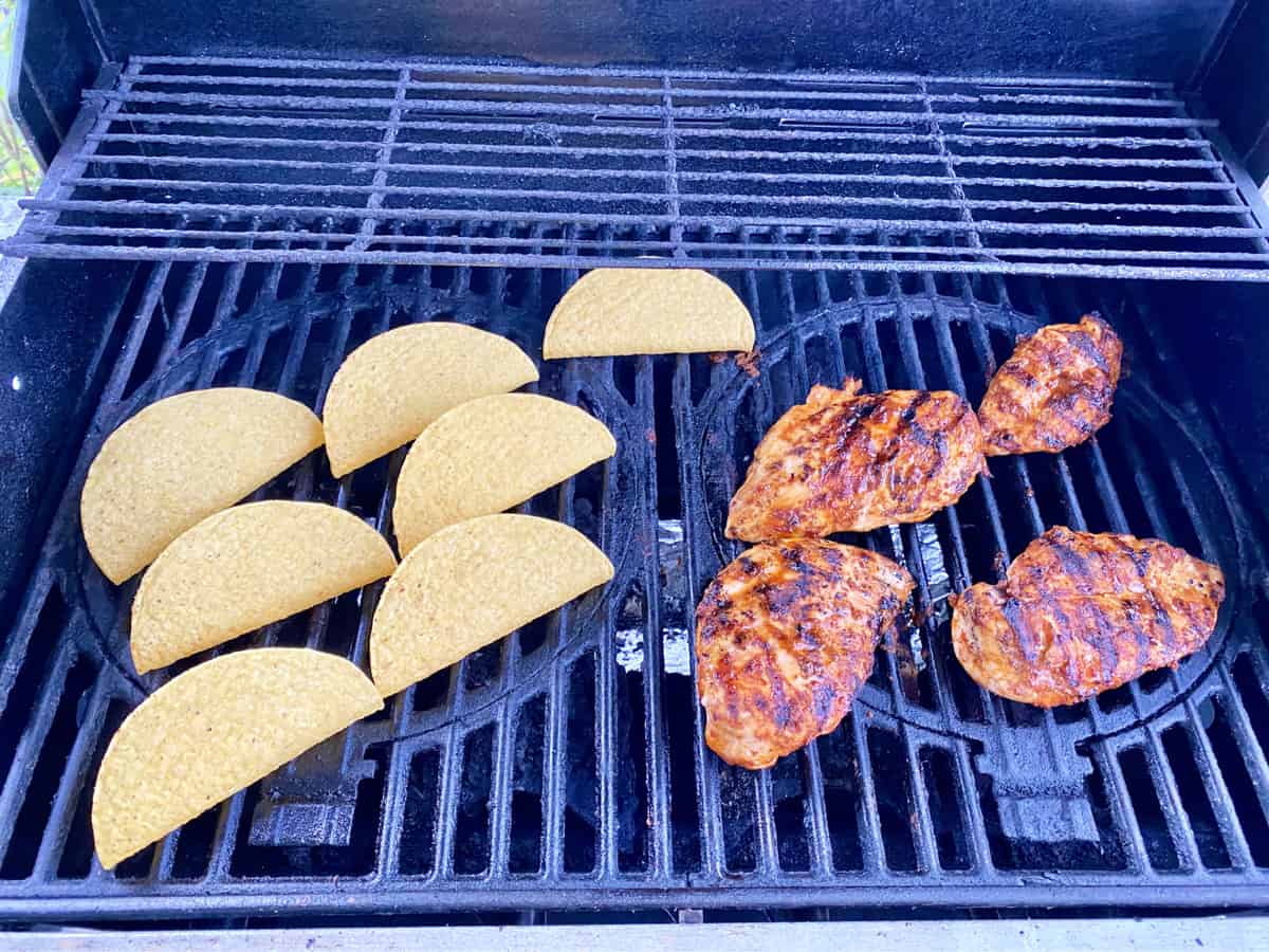 Grilled chicken breast and hard corn taco shells on top of a lit grill. 