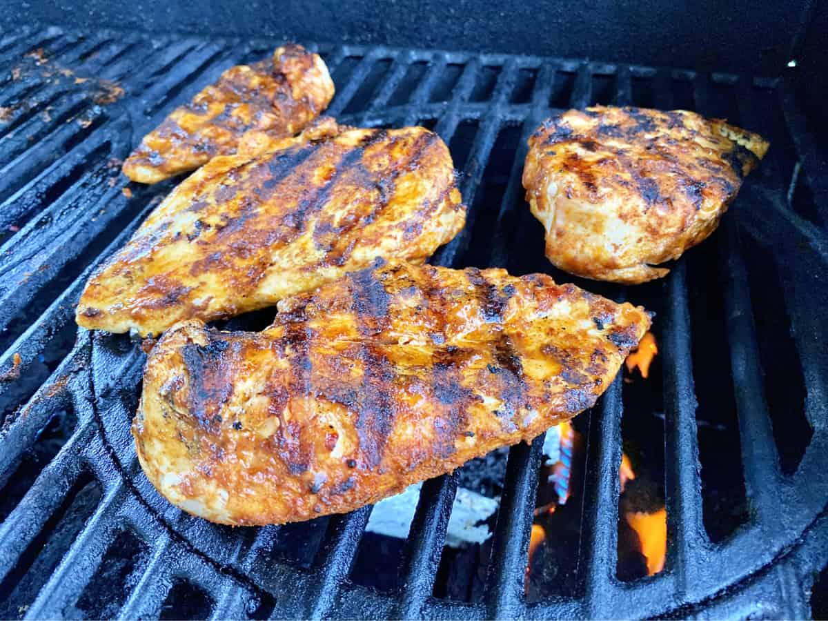 Four chicken breast with grill marks cooking on grill grates under orange fire. 