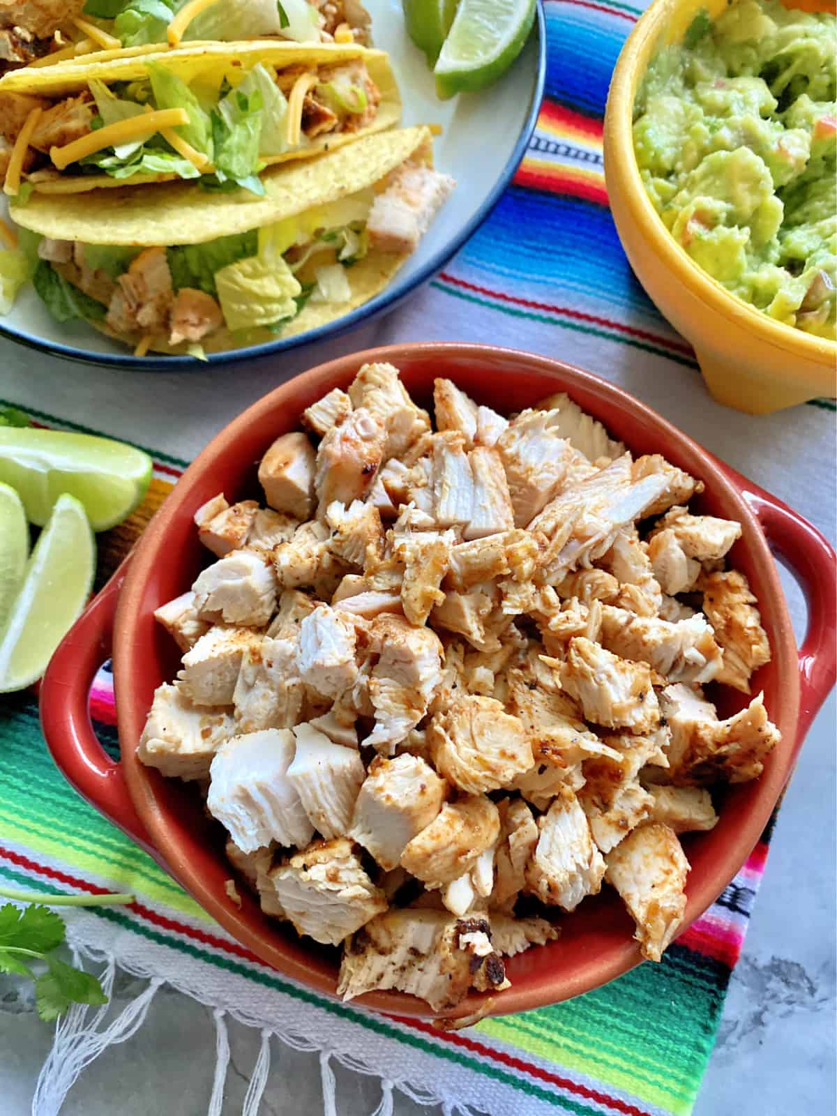 Top view of chopped chicken breast in a red bowl next to chicken tacos on a plate and guacamole. 