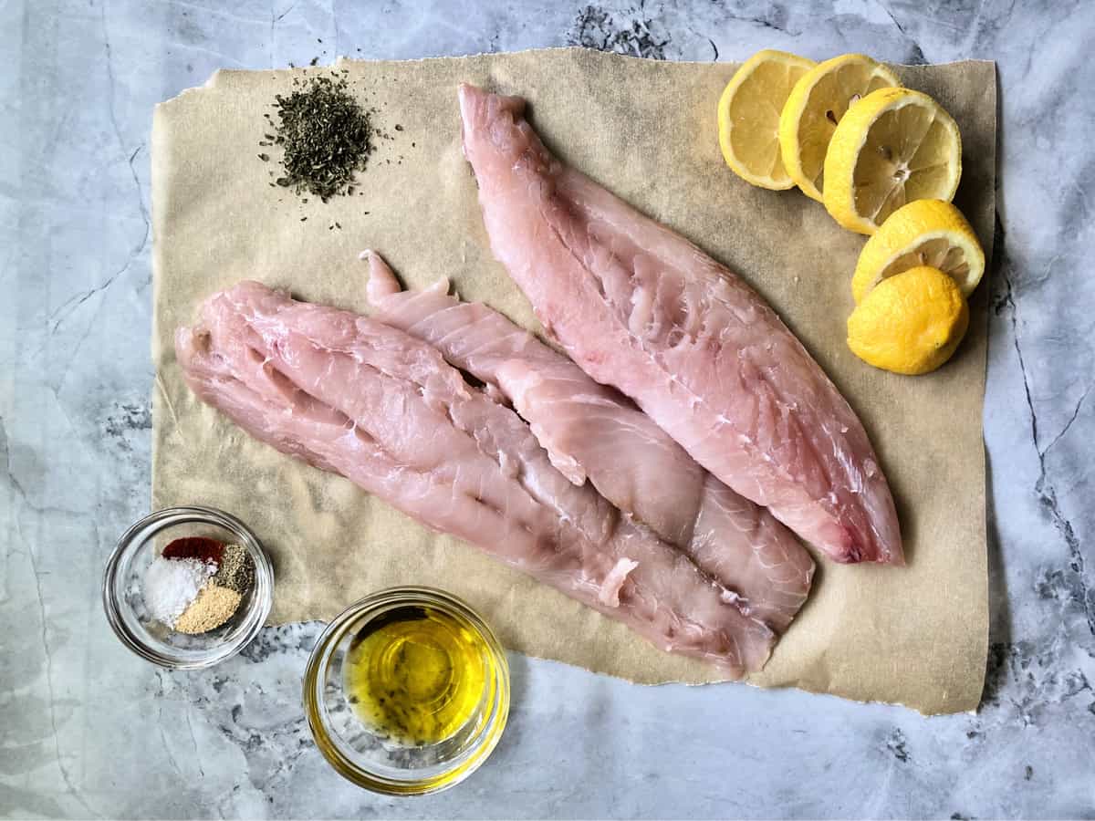 Two large slices of snapper on parchment paper with spices in a glass bowl, lemon, and olive oil.