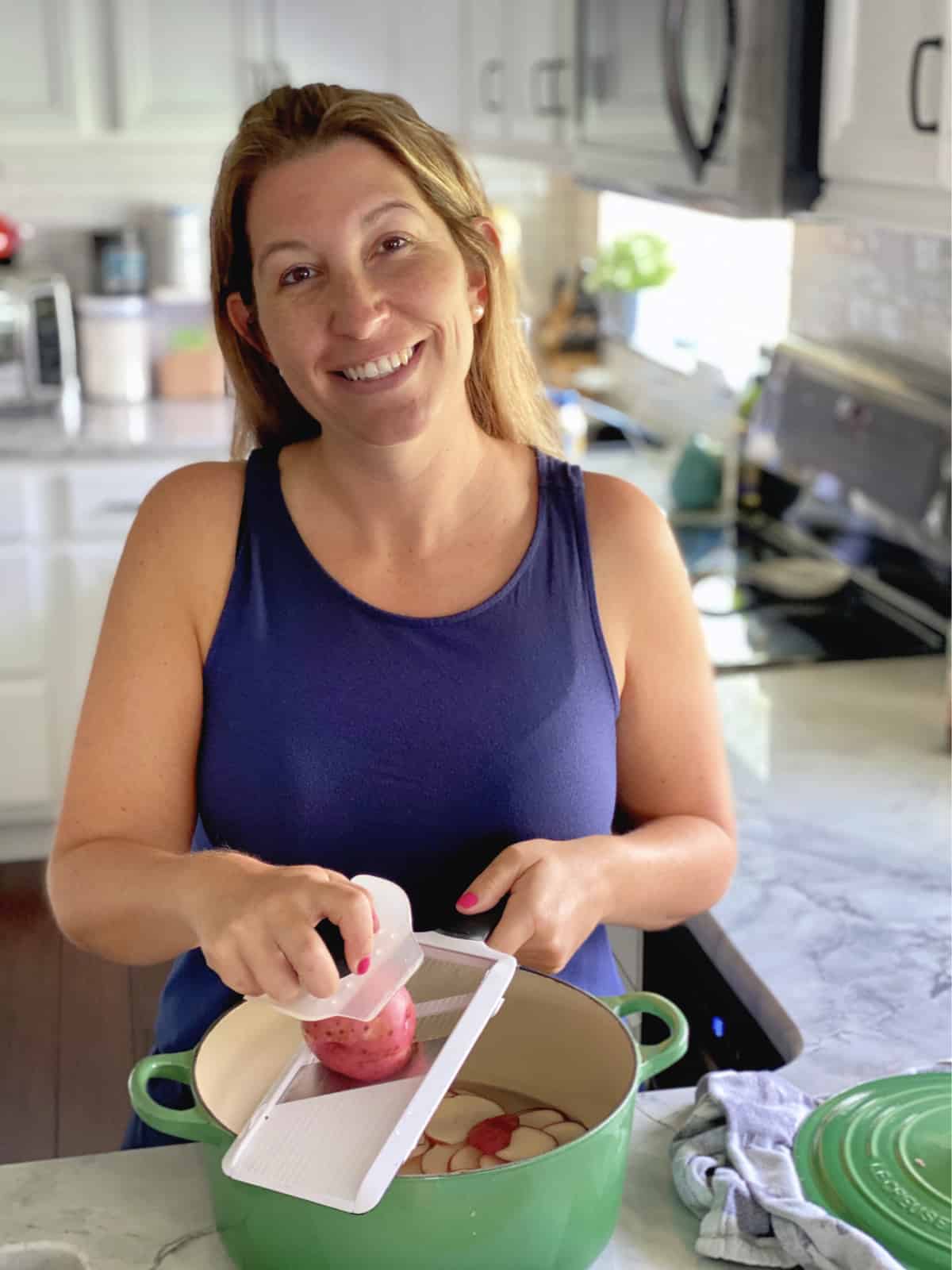 Female with brown hair and blue tank top in kitchen slicing potatoes into a green pot.