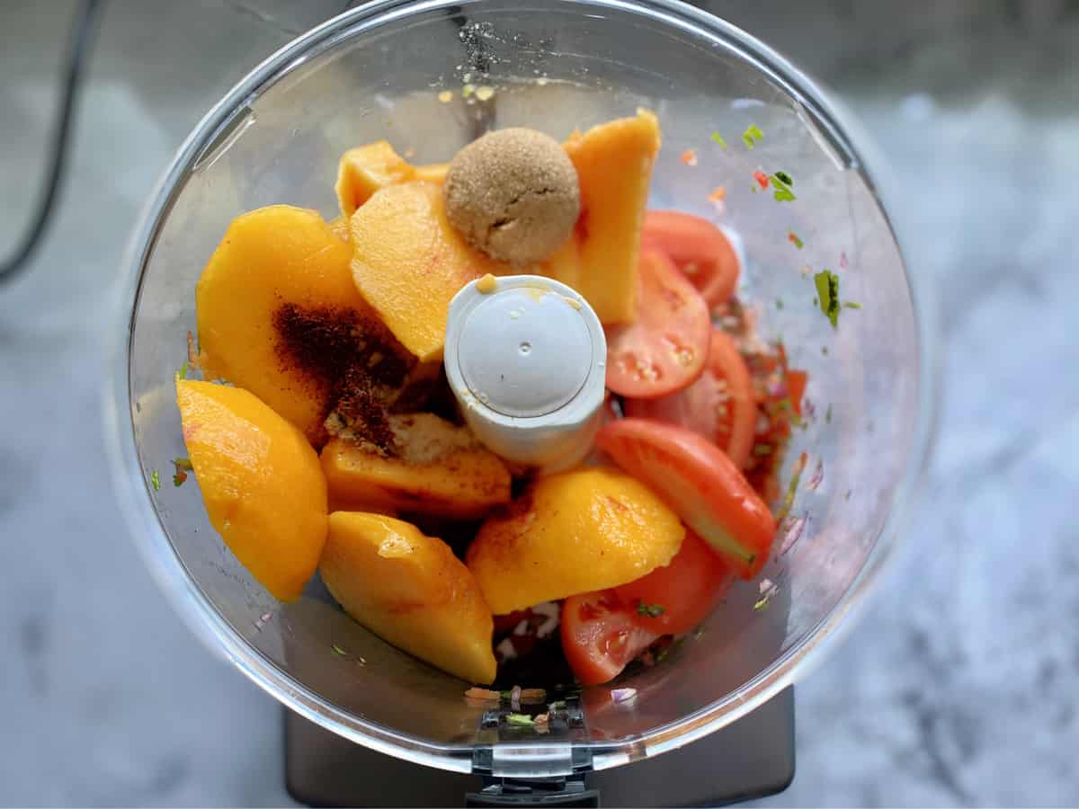 Top view of food processor with peach slices, spices, brown sugar, and plum tomatoes.