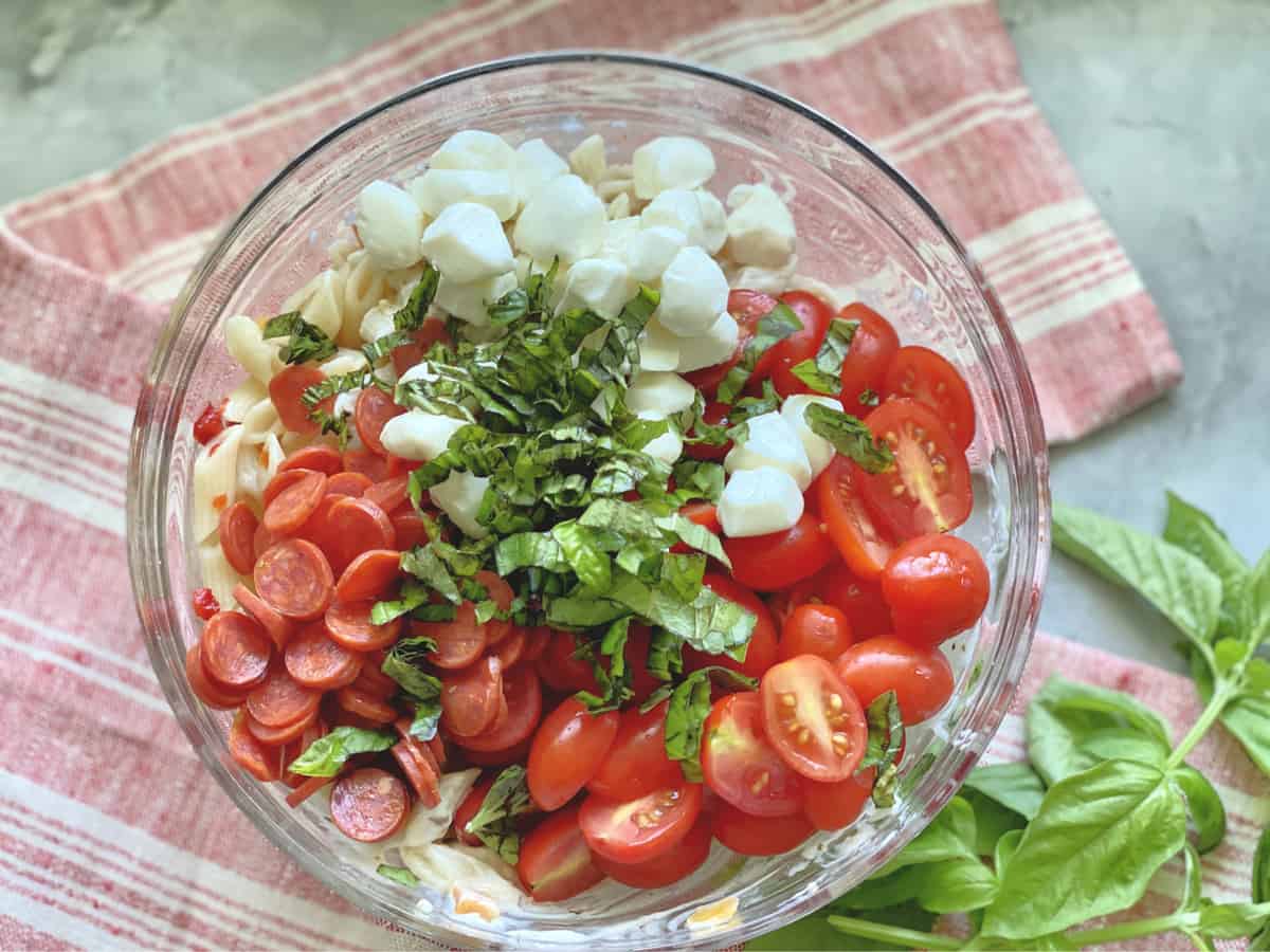 Top view of mini pepperoni, mozzarella pearls, sliced tomatoes and basil in a glass bowl.