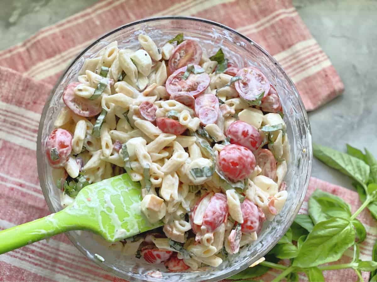 Mixed creamy penne pasta with sliced grape tomatoes and pepperoni in a glass bowl with green spatula.