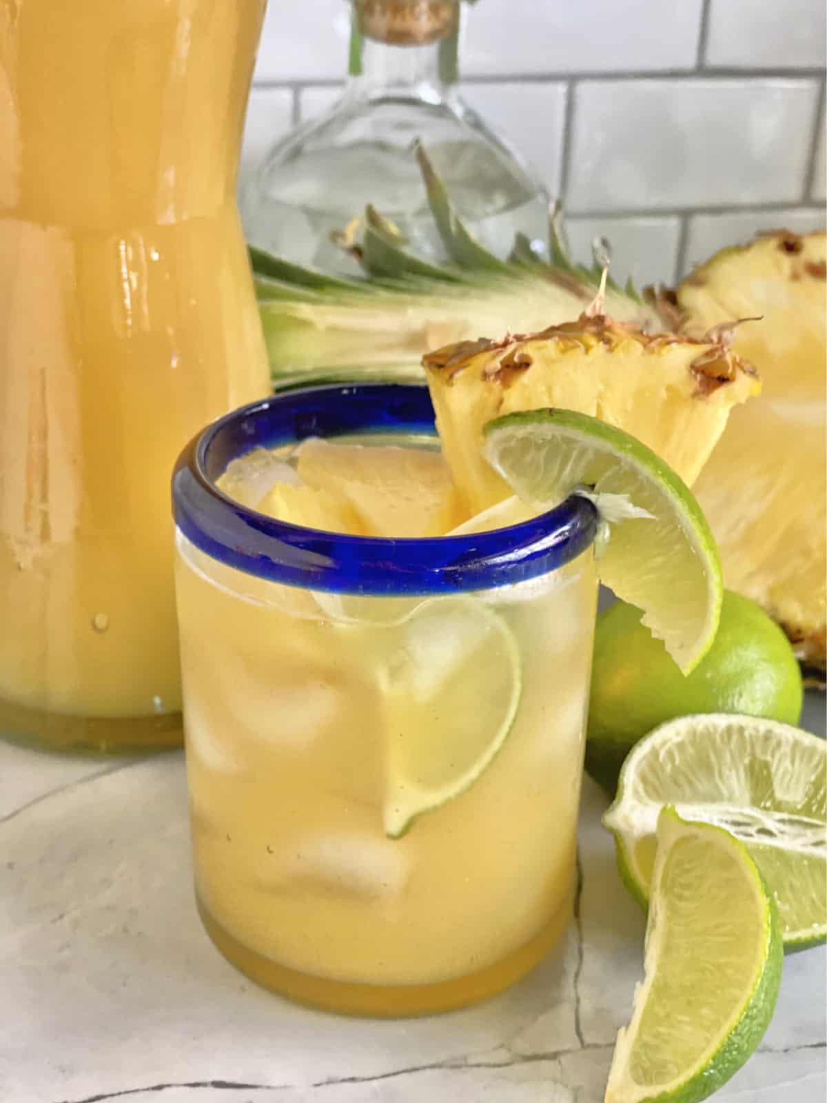 Close up of a glass with blue rim filled with a pineapple cocktail garnished with lime and pineapple.